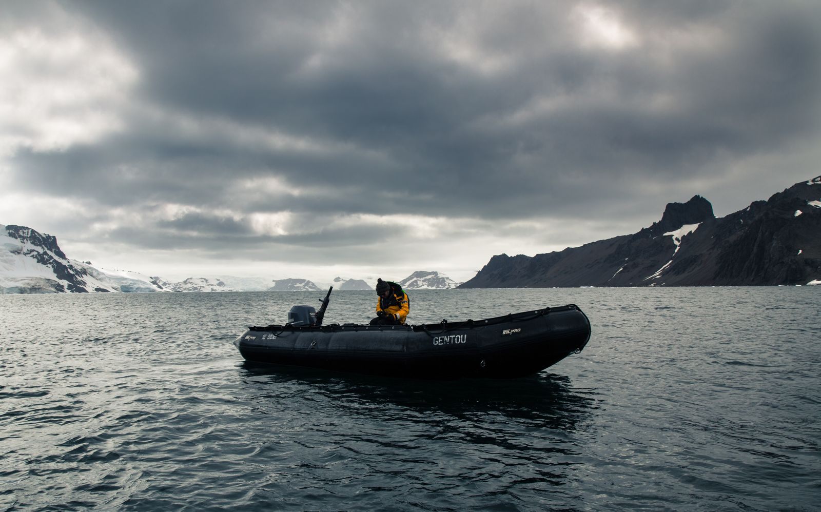 Inflatable expedition boat with a guide aboard on dark polar water below snowy ridges and heavy clouds