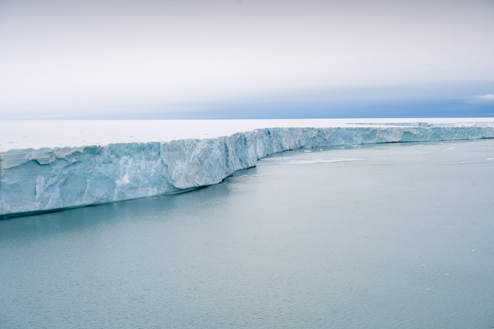 The towering blue-white cliff of an Antarctic ice shelf above calm sea, the scale that polar expedition cruises reach